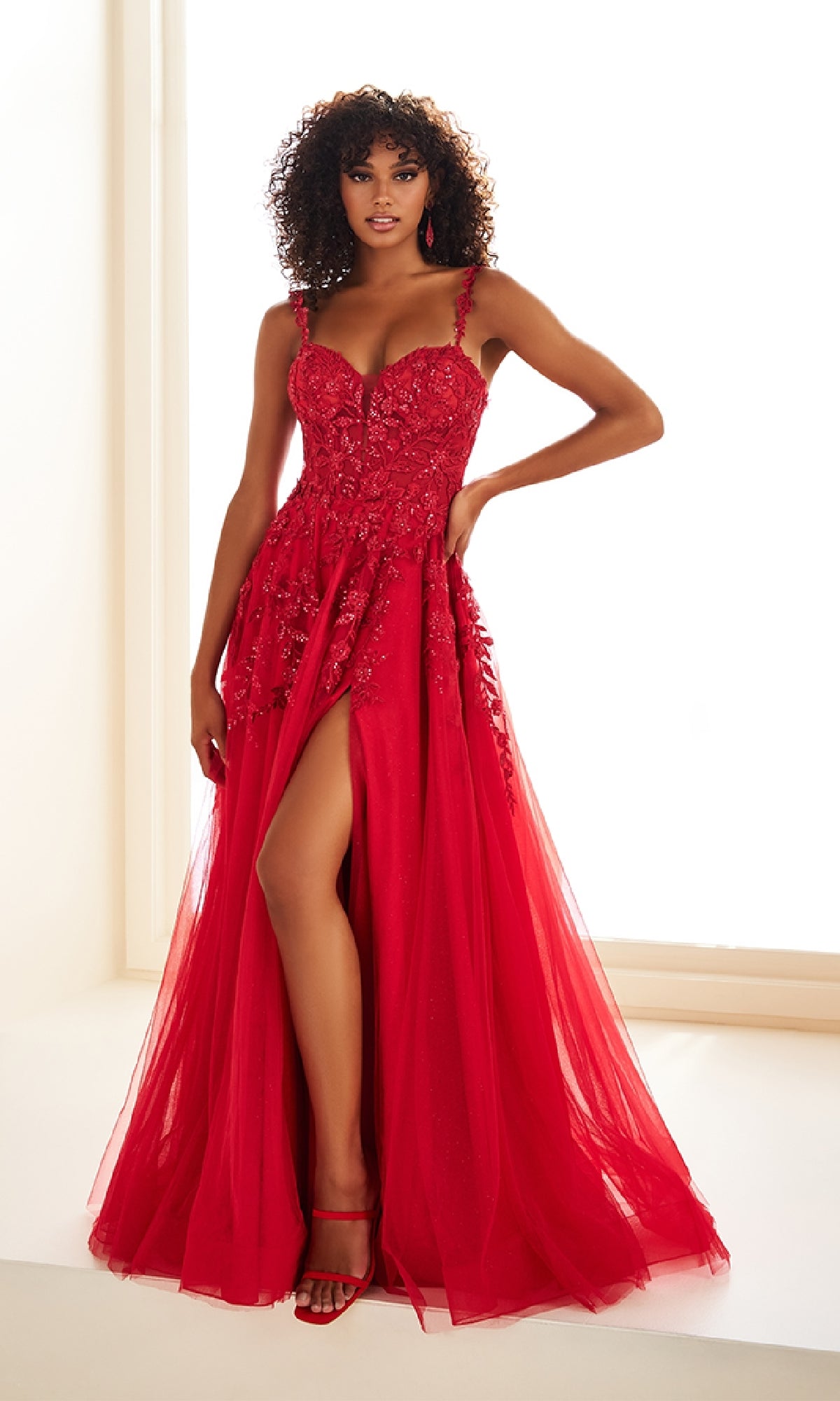A woman stands in front of a bright window wearing the Ellie Wilde EW36046 long prom dress—red, floor-length, with a high slit, floral embellishments, and spaghetti straps.